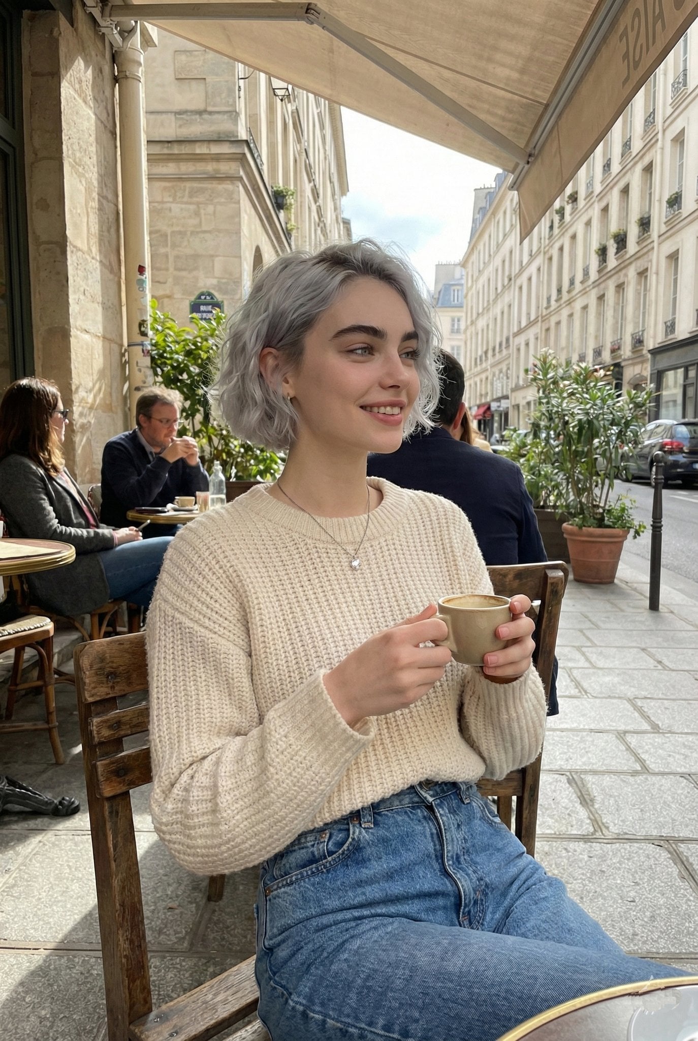 A candid lifestyle portrait of a young woman sitting at an outdoor Parisian café, holding a ceramic coffee cu…