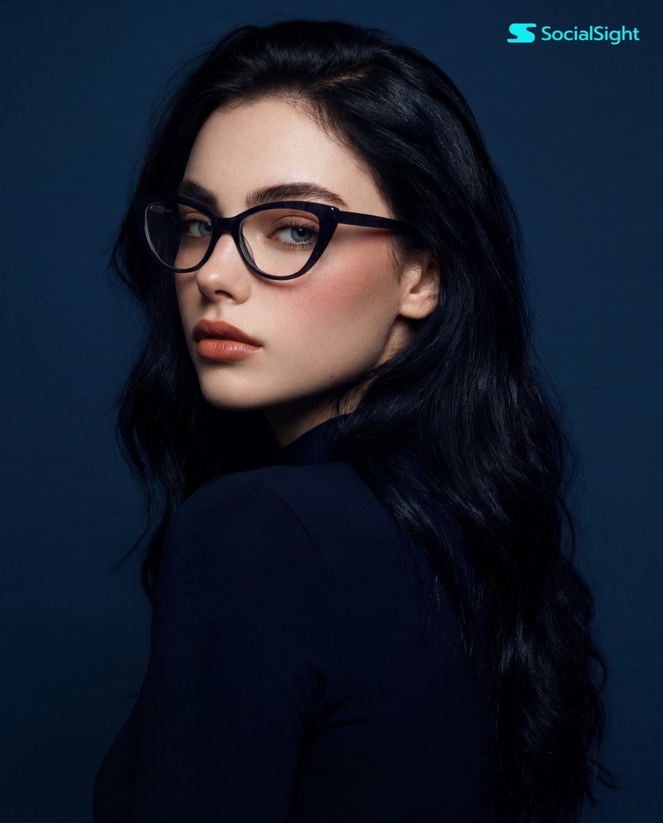 A young woman with long, dark hair and striking cat-eye glasses is captured in a studio portrait against a so…