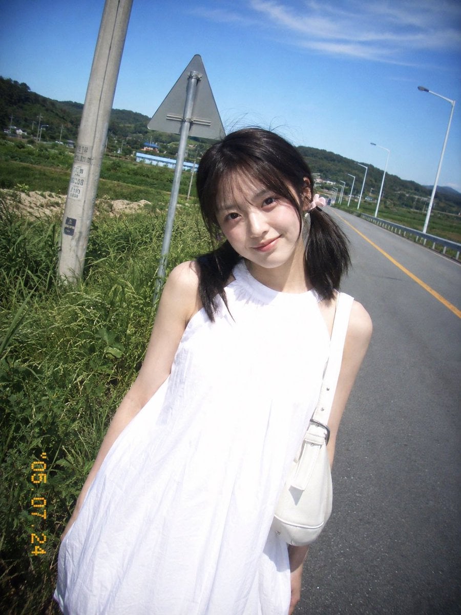 复古 CCD 卡片机夏日旅行抓拍，一位20岁出头的年轻东亚女idol站在乡间公路边。黑棕色低位双马尾，细碎刘海，浅粉色发圈，淡妆，冷白皮，…