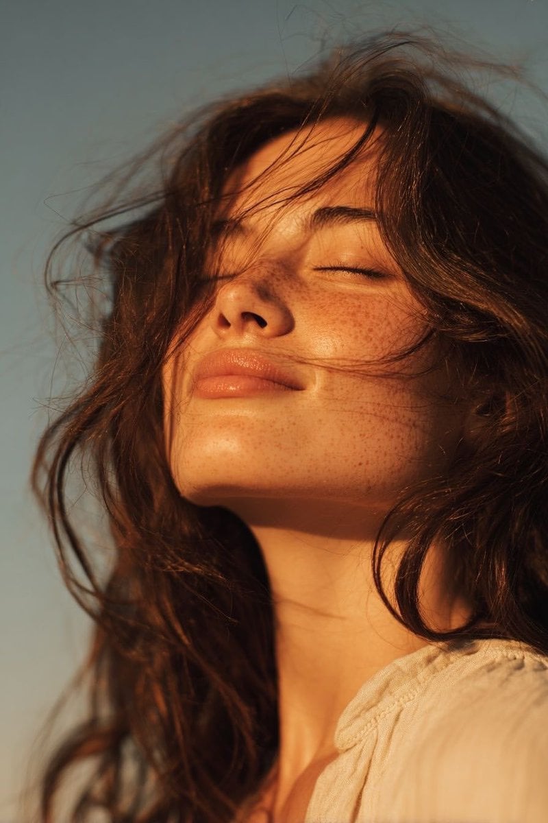 Ultra-realistic golden hour close-up portrait of a young woman outdoors, eyes gently closed with a soft peace…